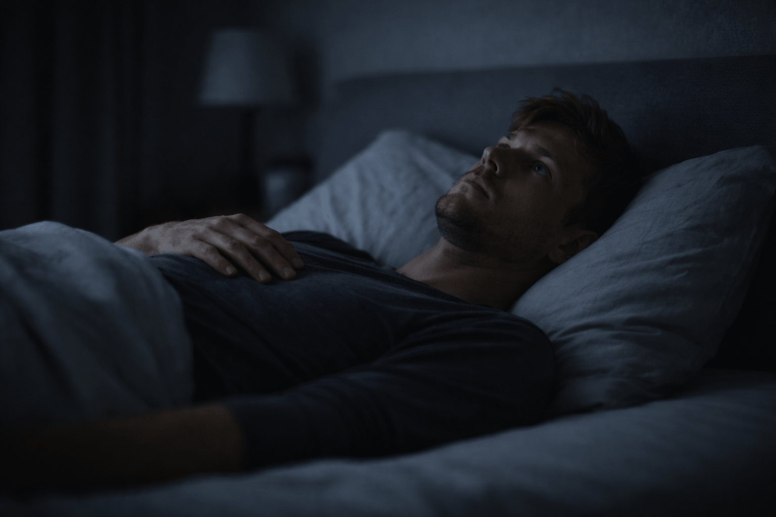 why do i feel guilty for resting – a person lying awake on a bed in a dark room, staring at the ceiling with subtle tension and inability to relax